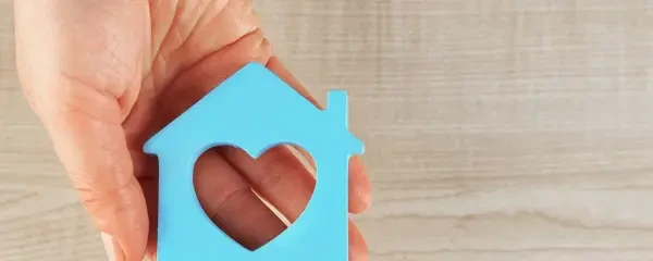 open hand holding a bright blue wooden flat shape of house with heart shape cut out